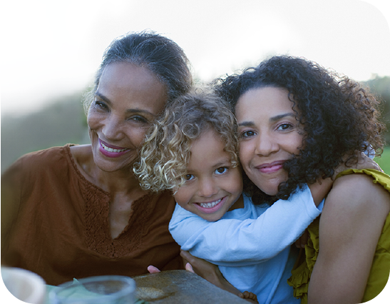 A mother, daughter, and young granddaughter smile toward the camera while hugging.